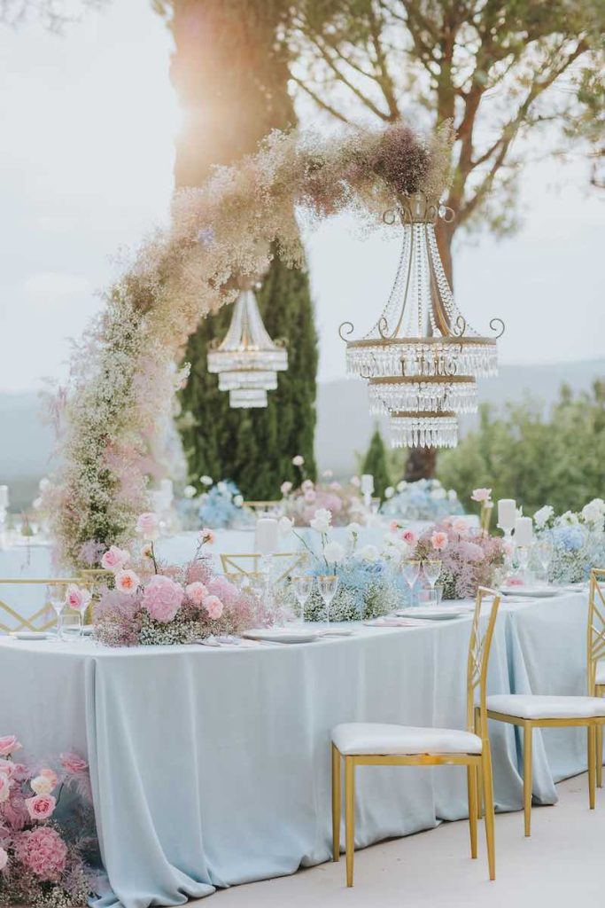 In questa foto l'allestimento di un matrimonio con gypsophila e rose nei toni dell'azzurro e del rosa. Sui tavoli imperiali pendono lampadari di cristallo e accanto sono posizionate sedie di colore oro