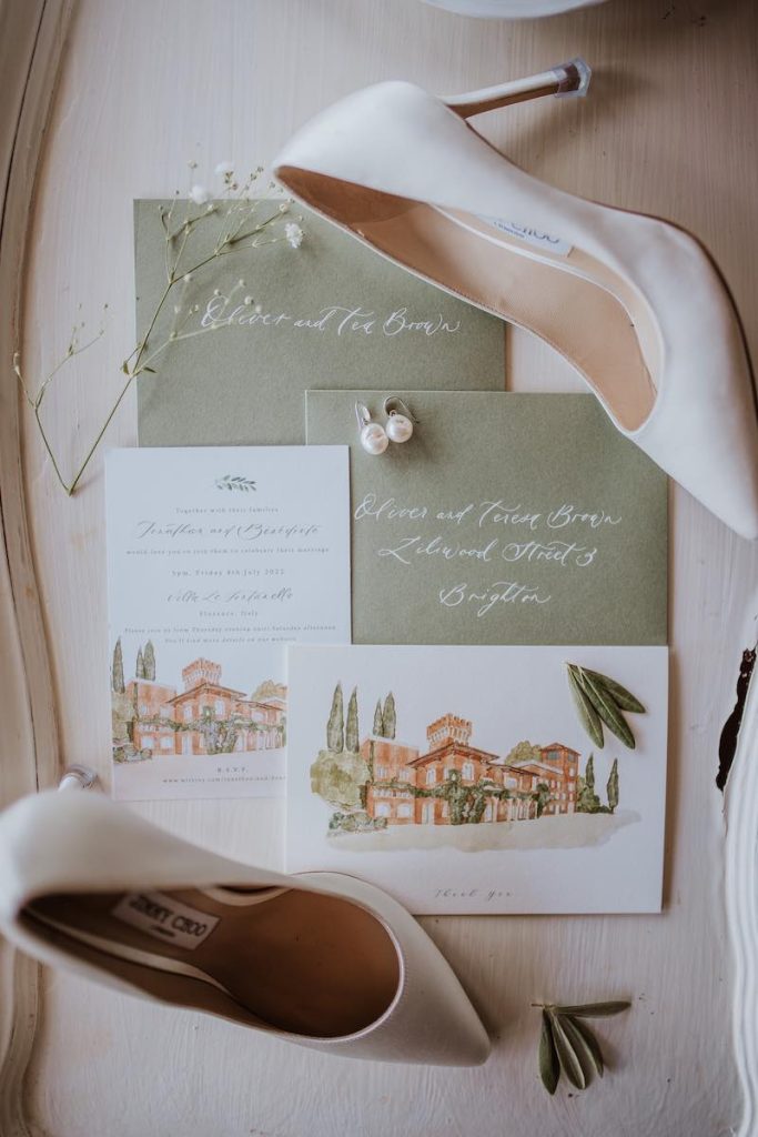 In questa foto una partecipazione di matrimonio nei toni del verde salvia e del bianco. Nel foglio di invito è riportato il disegno ad acquerello della location. Sulla partecipazioni sono posizionate le scarpe e gli orecchini della sposa