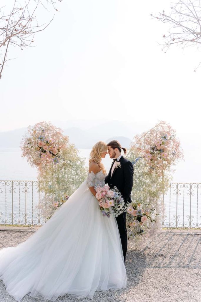 In questa foto due sposi con la testa poggiata l'uno all'altra davanti ad un backdrop a forma di ali composto da gypsophila e rosa nei toni pastello. La sposa tiene un bouquet con fiori abbinati