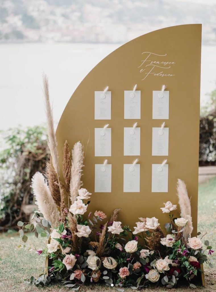 In questa foto un tableau de mariage a forma di mezza luna di colore oro con i biglietti con le liste dei tavoli di colore bianco. Davanti è presente una composizione con rose rosa e pampas