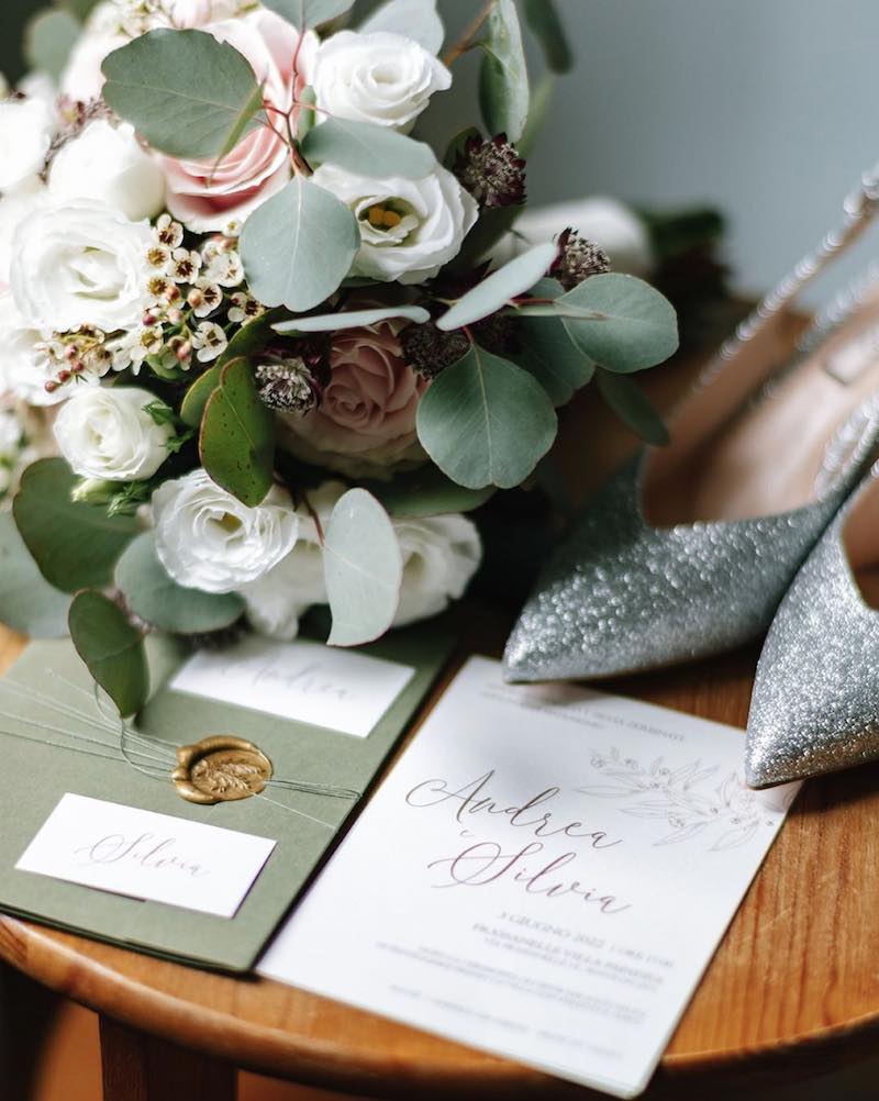 In questa foto una partecipazione di matrimonio nei toni del bianco, del verde e dell'oro. Dietro alla partecipazione sono posizionati il bouquet e la scarpe con glitter argento della sposa 