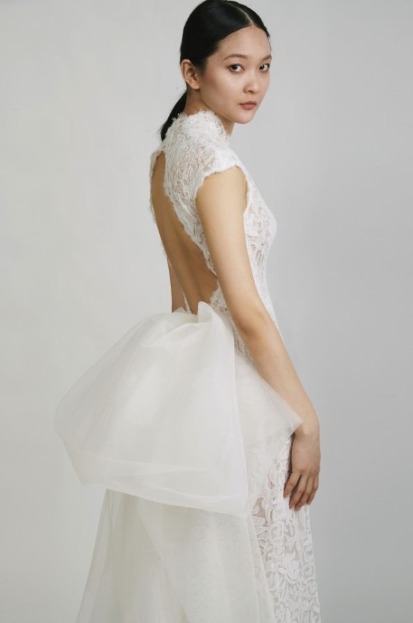 In questa foto una modella ritratta a mezzo busto indossa un abito Tosca Spose 2023 in pizzo con scollatura ad oblò sulla schiena e maxi fiocco in organza sul retro