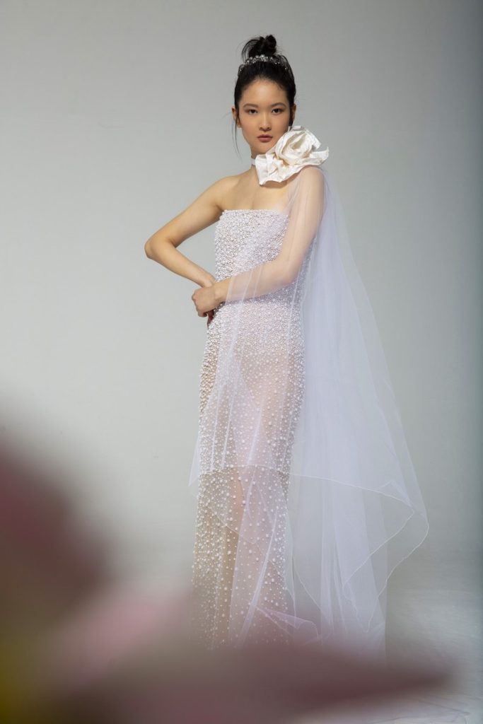 In questa foto una modella indossa un abito Tosca Spose 2023 nude con ricamo di perline all over con velo fermato sulla spalla sinistra da un maxi fiore color rosa pesca