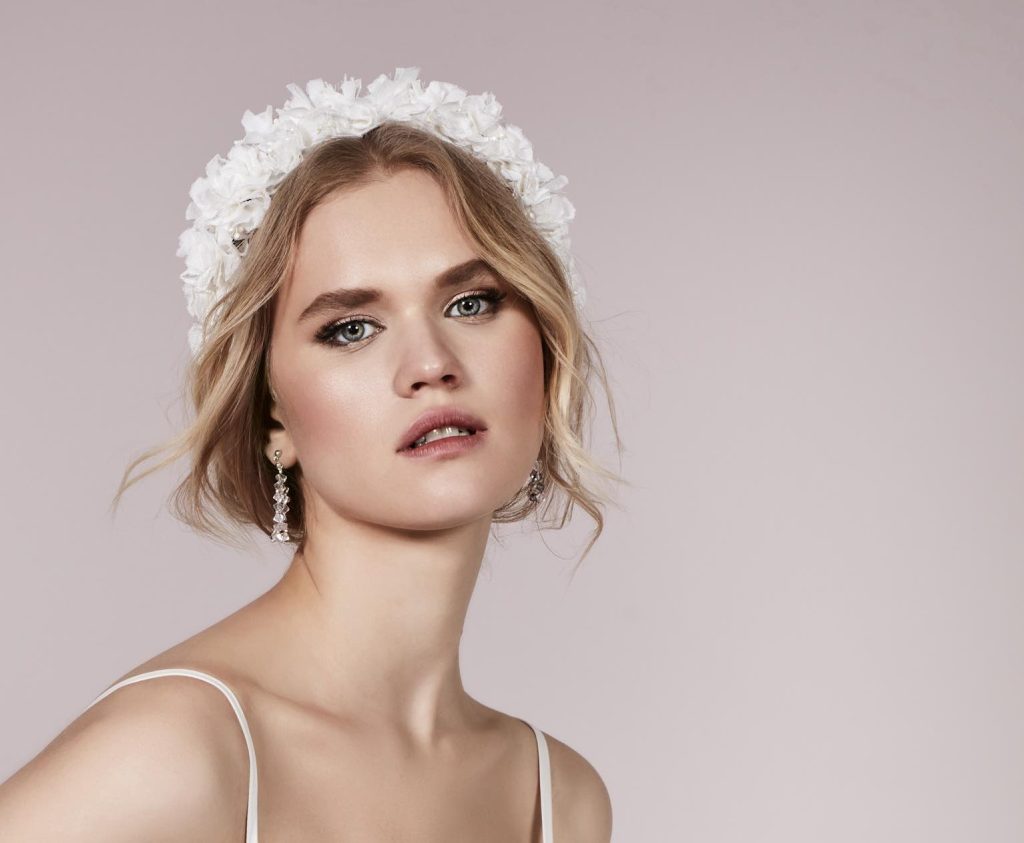 In questa foto una modella indossa un cerchietto con fiori di colore bianco e mostra un trucco sposa 2023 romantico