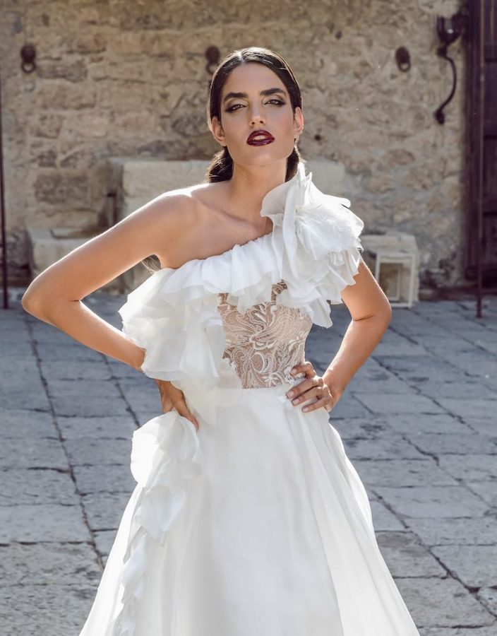 In questa foto una modella posa con un abito da sposa monospalla con ruches e bustino trasparente mostrando un trucco sposa vintage con rossetto color prugna lucido