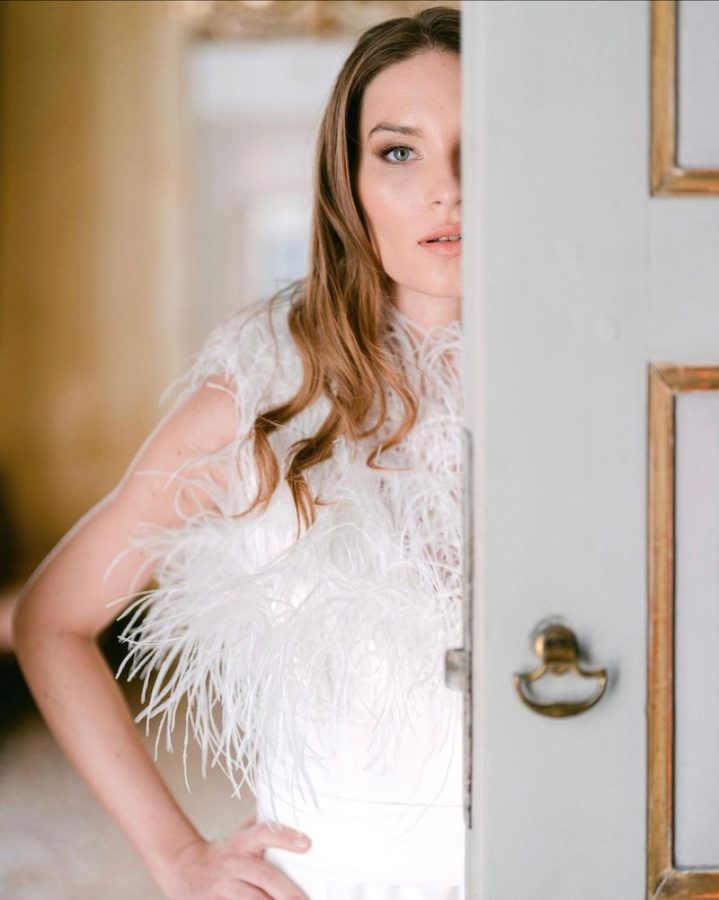 In questa foto una modella posa dietro una porta di un palazzo antico, indossa un abito da sposa con piume sul corpetto e un trucco sposa nude 2023