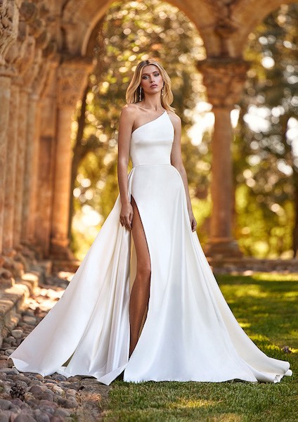 In questa foto una modella indossa un abito da sposa monospalla in bianco ottico e con profondo spacco. Il vestito è di Pronovias 2023