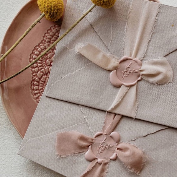 In questa foto una busta chiusa da un fiocco realizzato con un nastro in seta rosa cipria, fissato sulla carta da un sigillo in ceralacca ton sur ton
