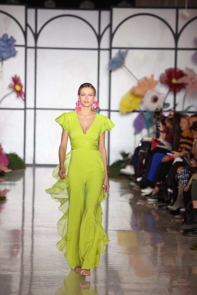 In questa foto una modella sfila indossando una tuta di colore verde lime della collezione cerimonia Atelier Emé 2023. La tuta presenza uno scollo con maniche corte con ruches 