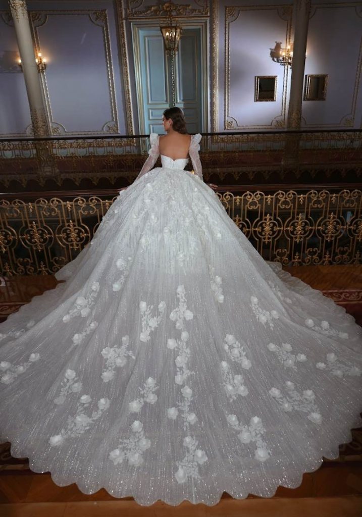 In questa foto una modella indossa un abito da sposa Dovita Bridal 2023 con lunghissimo strascico ricamato con glitter e pizzo rebrodé 