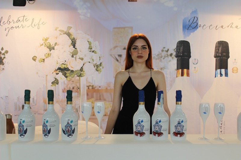 In questa foto una modella dietro ad un banco sui cui sono disposte sei bottiglie Bocciamatta accompagnati da calici Bocciamatta