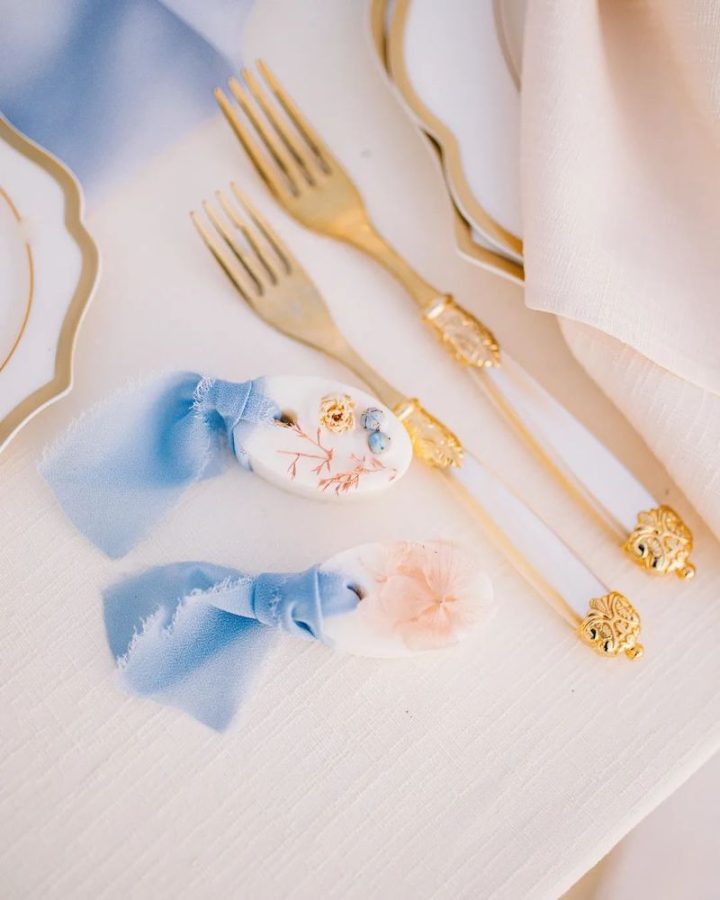 In questa foto duce cialde di cera di soia con fiori essiccati decorate con nastri di colore azzurro e poggiate su un tavolo di nozze accanto a due forchette dorate e scelte come bomboniere matrimonio 2023
