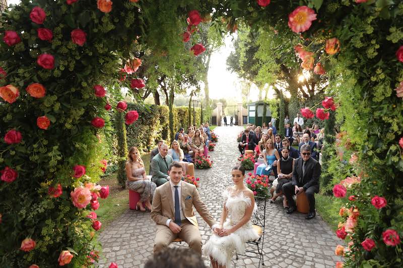 In questa foto i due sposi seduti davanti all'arco di fiori di colore fucsia e arancioni nel giardino di una villa. Dietro agli sposi sono seduti tutti gli ospiti 