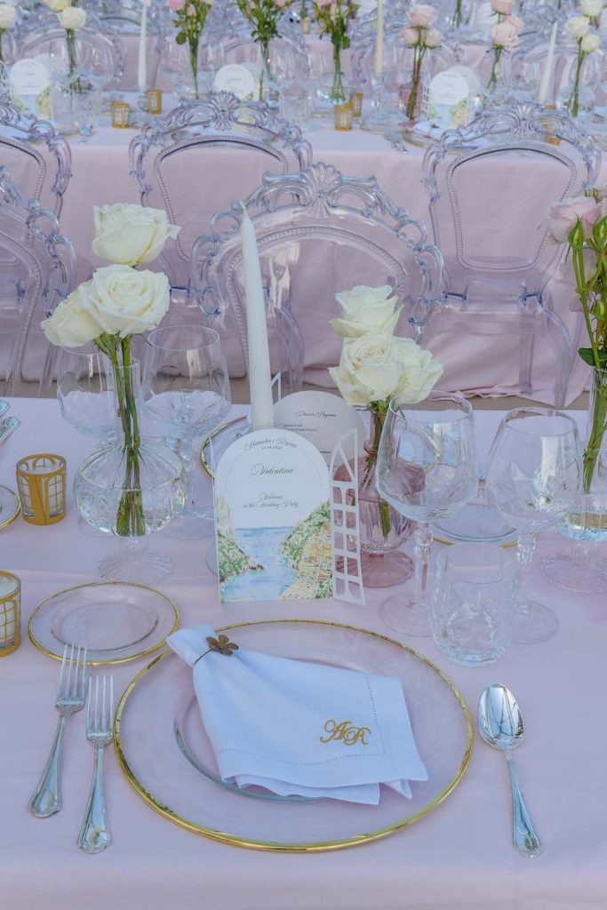 In questa foto una mise en place di Cira Lombardo con tovagliato rosa blush, piatti trasparente con bordi oro, rose bianche in vasetti trasparenti, segnaposto intagliati con una vista della Costiera Amalfitana