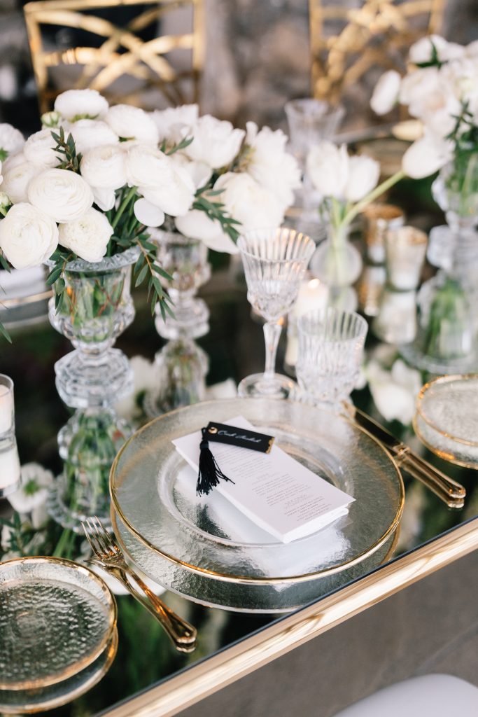 In questa immagine, una mise en place matrimonio 2023 con piatto con bordo in oro, bicchieri di cristallo e vasi ripieni di peonie fresche. 