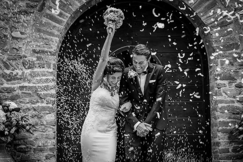 In questa foto in bianco e nero di Nicola Tanzella due sposi escono dalla chiesa durante il lancio di riso e petali. La sposa alza la mano trionfante con il bouquet