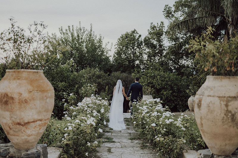 In questa foto dello Studio Santanastasio due sposi di spalle camminano in una stradina di ciottoli circondata da fiori bianchi e vasi in terracotta 