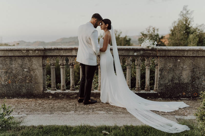 In questa foto dello Studio Santanastasio due sposi davanti ad una balaustra in marmo si appoggiano ad occhi chiusi l'uno sulla fronte dell'altra. La sposa indossa un abito a sirena liscio e un velo a taglio vivo. Lo sposo indossa uno smoking con giacca bianca