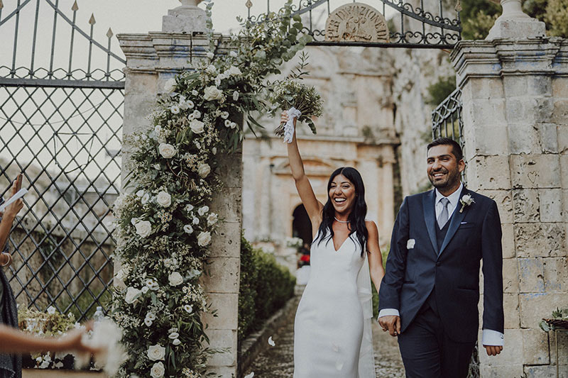 In questa foto dello Studio Santanastasio due sposi sorridenti dopo la cerimonia in chiesa passano attraverso un arco decorato con fiori bianchi. La sposa alza trionfante con la mano destra il suo bouquet