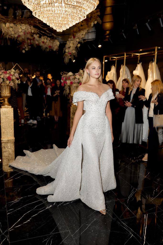 In questa foto una modella sfila nell'atelier di Milano con uno degli abiti della collezione premium Time Dovita Bridal. Si tratta di un abito a sirena con sovragonna, scollatura off shoulder, tessuto sparkling e ricami di perline