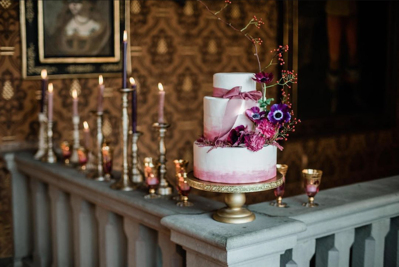 In questa foto torta per matrimonio 2023 a 3 piani con effetto watercolor di colore porpora sulla base di ogni piano. La decorazione è completata con un nastro di colore rosa e fiori abbinati. La torta è posizionata su una balaustra di marmo decorata con portacandele di colore oro e candele rosa