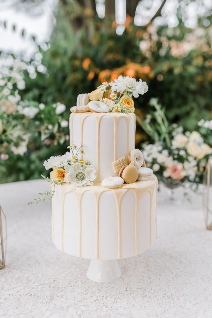 In questa foto una drip cake di colore bianco e panna decorata con fiori, ciambelline e macarons