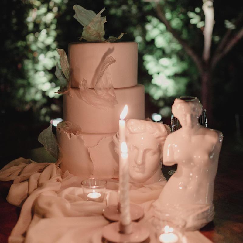 In questa foto una torn paper cake a piani di colore bianco posizionata su tavolo al buio e circondata da da statuette di porcellana e candele accese