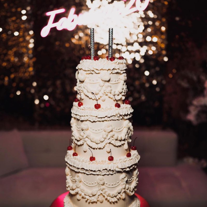 In questa foto una Lambeth Cake per matrimonio decorata con glassa di colore bianco e ciliegine rosse glassate. Sullo sfondo si intravede una scritta al neon