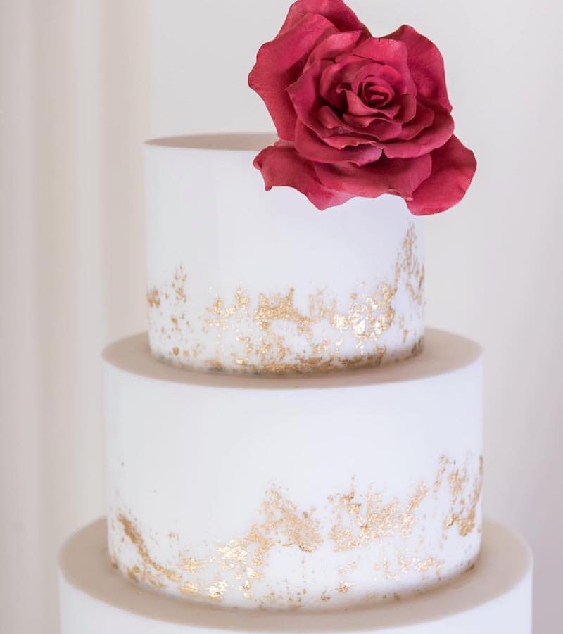 In questa foto una torta matrimonio a piani decorata con glassa di colore bianco, screziature d'oro e una rosa di colore rosso usata come cake topper