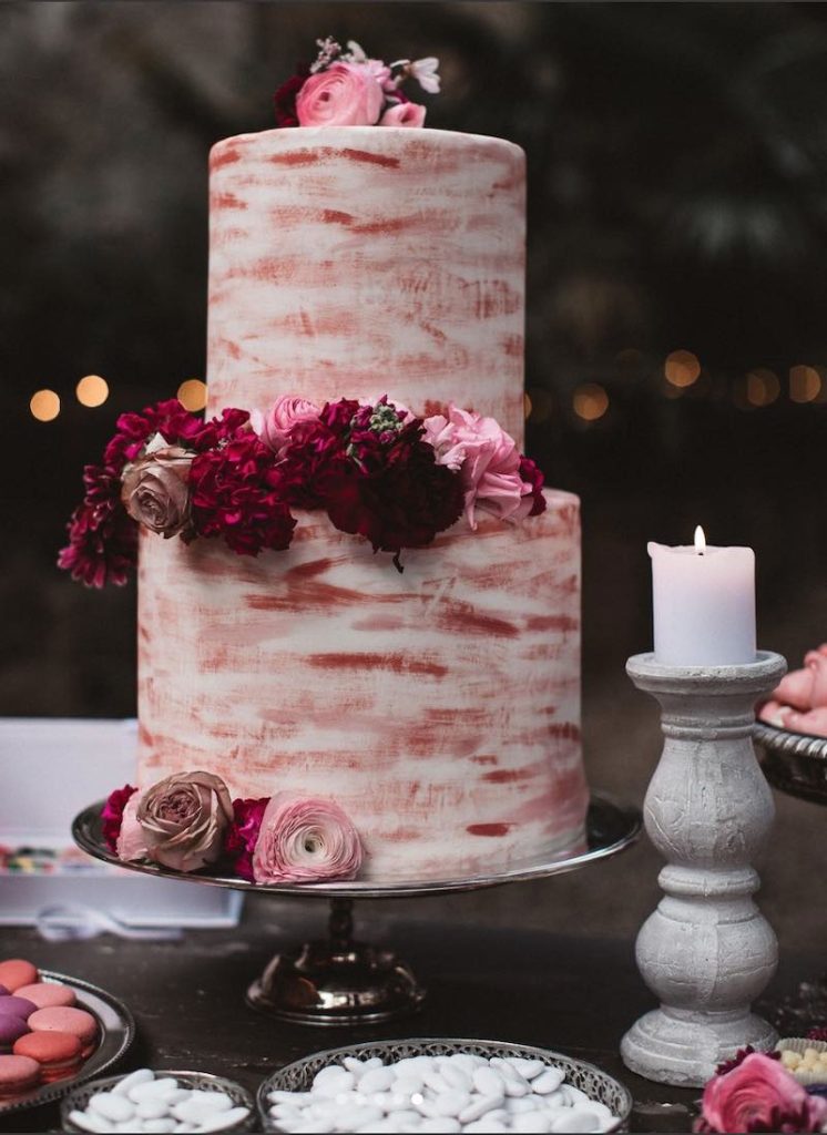 In questa foto una torta nuziale decorata con glassa di colore bianco e pennellate di colore rosso e fiori abbinati. La torta è posizionata su un piatto d'argento circordata da candele e portaconfetti
