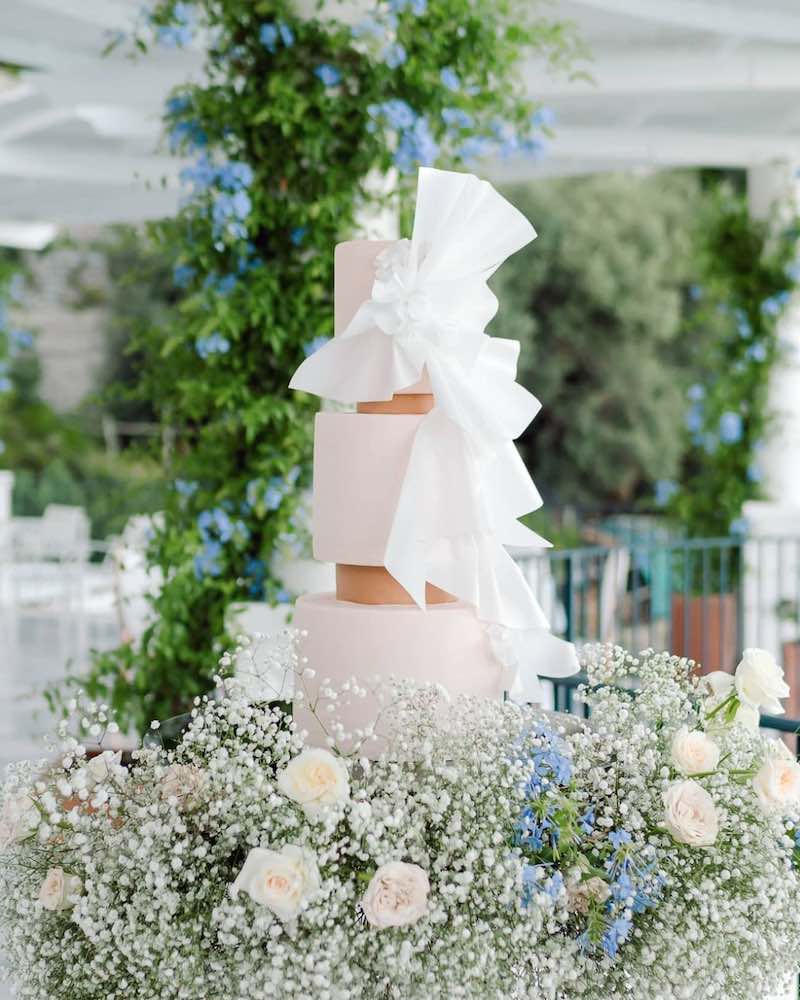 In questa foto una torta per matrimonio a piani separati in bianco e oro decorato con un maxi fiocco di colore bianco. La torta è posizionata tra gypsophila di colore bianco e rose di colore bianco e azzurro