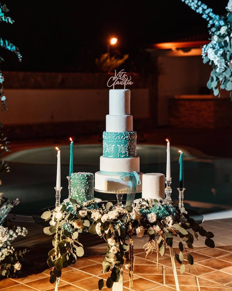 In questa foto una torta per matrimonio a 5 piani decorata con glassa di colore bianco e a metà con glassa di colore verde tiffany e pizzi. In cima è presente un. cake topper con i nomi degli sposi a taglio laser. La torta è posizionata su un tavolo decorata con foglie e rose di colore bianco e candele accese