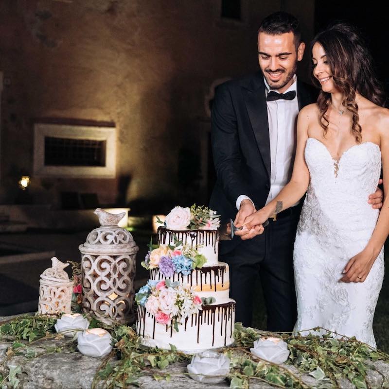 In questa foto due sposi tagliano una drip cake per matrimonio con cioccolato e decorata con fiori multicolor
