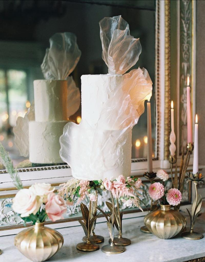 In questa foto una delle torte matrimonio 2023 decorata con glassa di colore bianco e fogli di pasta di riso trasparente. La torta è posizionata su un piano di marmo davanti ad uno specchio ed è circondata da vasetti di colore oro con fiori rosa 