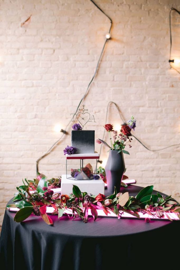 In questa foto una torta per matrimonio a piani separati. Il primo piano è decorato a watercolor nero e bianco; il secondo è un ottagono di vetro; il terzo è di colore bianco. La torta è poggiata su un tavolo rotondo davanti ad un muro industrial di colore bianco