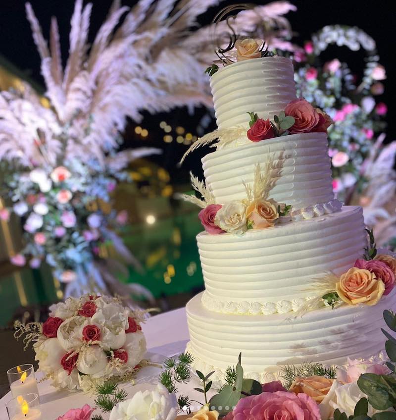 In questa foto una delle torte matrimonio 2023 a 4 piani decorata con glassa bianca e fiori di colore giallo e rosa. Sullo sfondo si intravede un allestimento floreale con rose e pampas