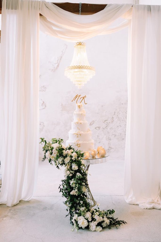In questa foto una torta per matrimonio a 5 piani decorata con glassa e fiorellini di pasta di zucchero di colore bianco. È poggiata su un tavolo allestito con un cordono di foglie e fiori di colore bianco e sotto un lampadario di cristallo