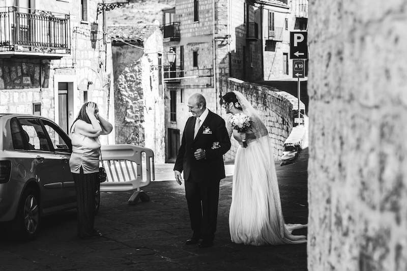 In questa foto in bianco e nero di Vincenzo Aluia una sposa al braccio del papà sorride ad una signora con le mani sulla testa incontrata per le strade di Petralia Sottana