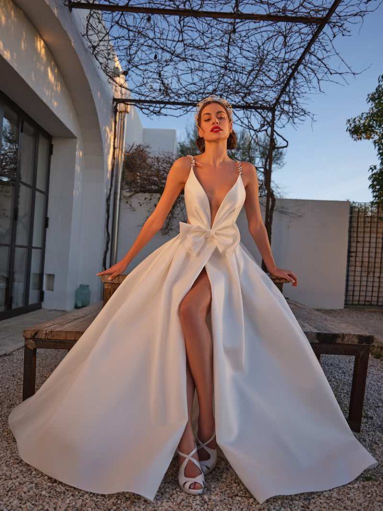 In questa foto una modella indossa un abito da sposa Mysecret sposa 2024 della linea Orchidea modello ad A in mikado di seta con spacco frontale, fiocco in vita e scollo a V profondo. L'abito presenta anche spalline sottili gioiello