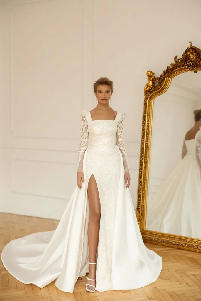 In questa foto una modella indossa uno degli abiti da sposa Eva Lendel 2023 a sirena con spacco alto fino alla coscia, scollo quadrato su corpetto drappeggiato, maniche lunghe in picco chantilly ripreso anche sulle lunghezze. L'abito è un modello trasformabile con sopragonna in mikado di seta 