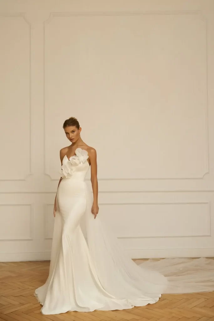 In questa foto una modella indossa un abito da sposa a sirena Eva Lendel 2023 in crêpe con sopragonna staccabile e maxi fiori 3D lungo la scollatura