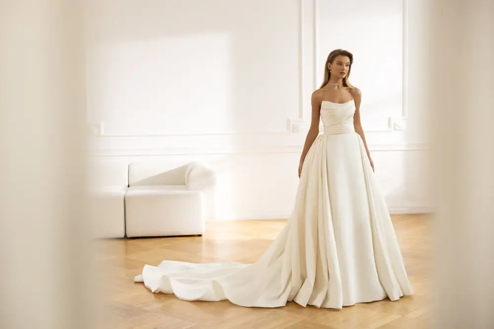 In questa foto una modella indossa uno degli abiti da sposa Eva Lendel 2023 trasformabili: è un modello a sirena in mikado di seta con corpetto drappeggiato e sopragonna staccabile