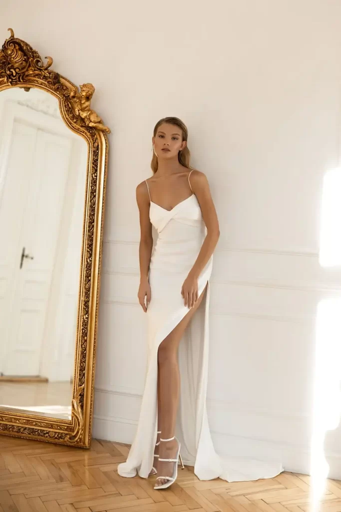 In questa foto una modella indossa un abito da sposa Eva Lendel 2023 a sirena modello lingerie con lungo spacco fino alla coscia. La modella indossa anche sandali abbinati alla schiava di colore bianco