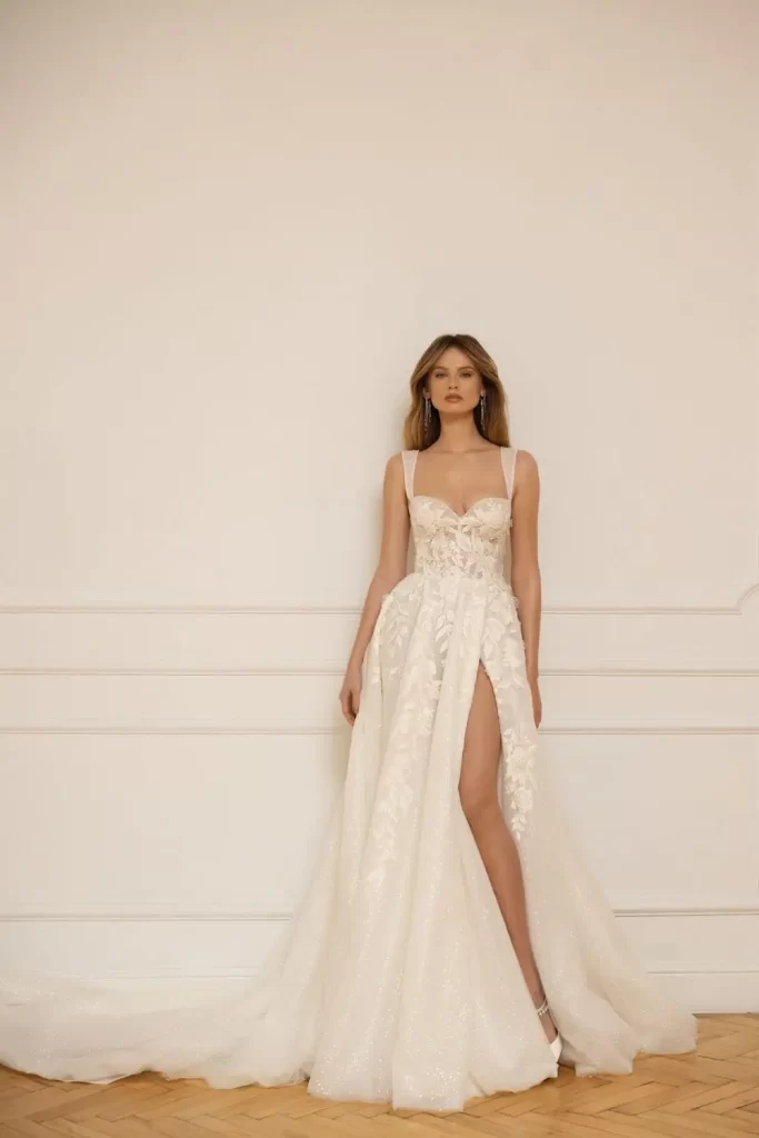 In questa foto una modella indossa uno degli abiti da sposa Eva Lendel 2023 modello scivolato in tulle ricamato in pizzo rebrodé con spacco e scollo a cuore con spalline larghe