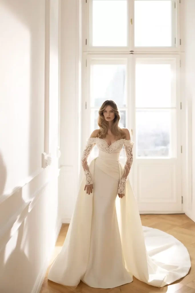 In questa foto una modella indossa un abito da sposa trasformabile Eva Lendel 2023 modello a sirena in mikado di seta con scollo off shoulder, maniche lunghe in pizzo e sopragonna in mikado di seta