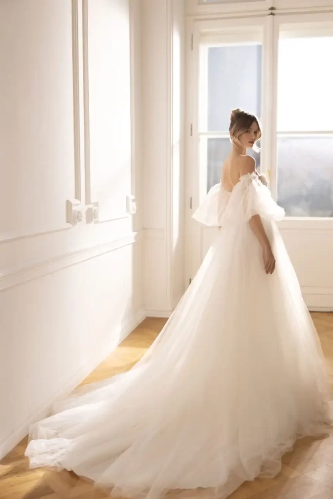 In questa foto una modella inquadrata di spalle indossa un abito da sposa Eva Lendel 2023 ampio con lungo strascico in tulle con maniche balloon 