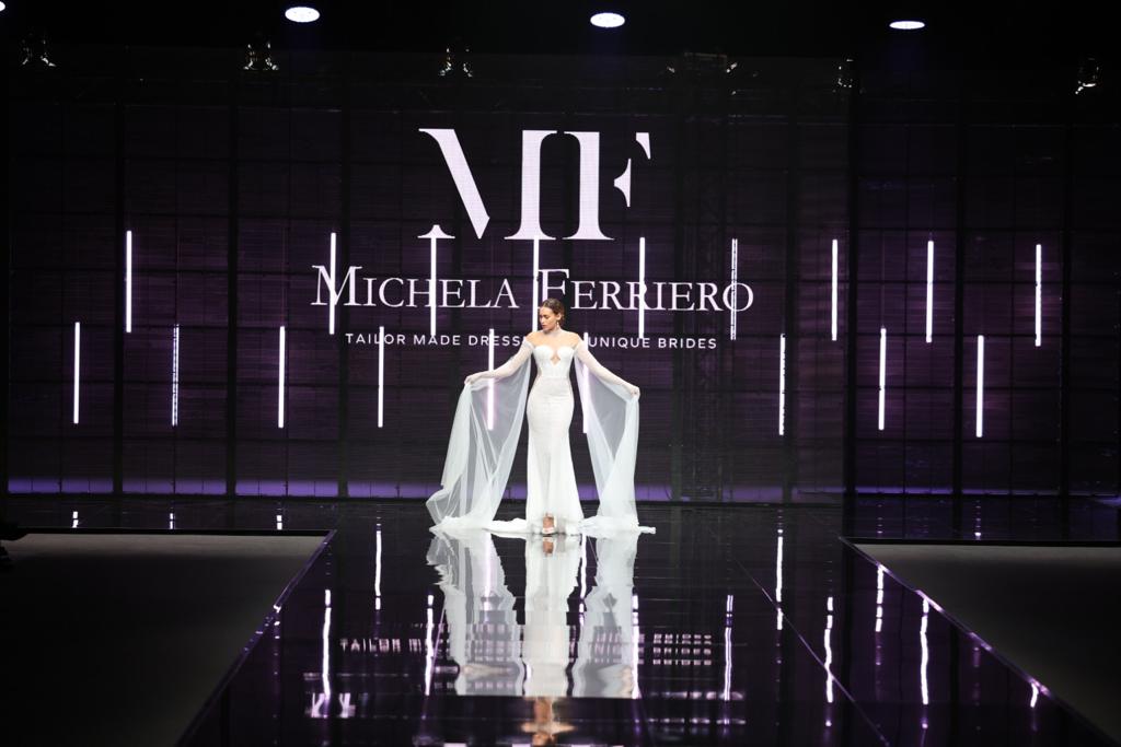 In questa foto una modella sfila indossando un abito da sposa Michela Ferriero 2024 a sirena con mani in chiffon ampie che si aprono come ali