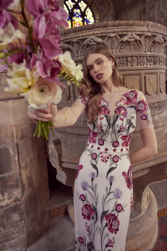 In questa foto una modella indossa un abito da sposa Temperley Bridal 2024 con ricami floreali di colore bordeaux, rosa e lilla. L'abito presenta maniche corte in tulle trasparente
