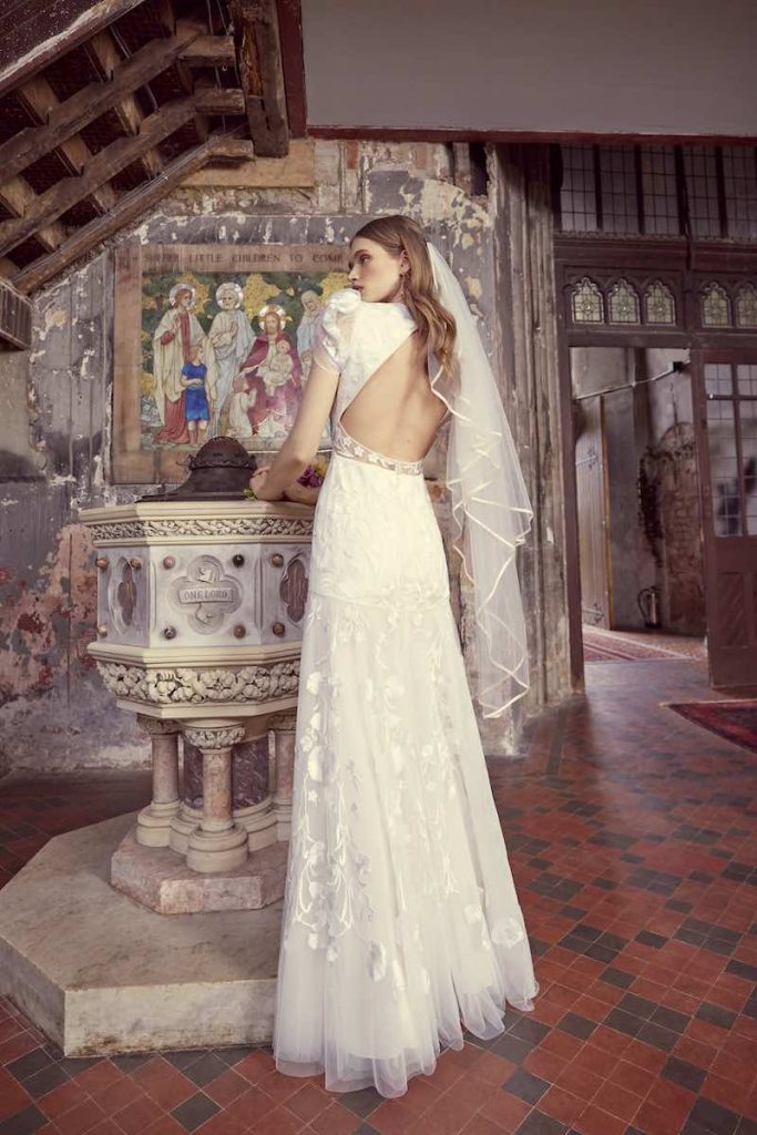 In questa foto una modella inquadrata di spalle indossa un abito da sposa Temperley Bridal 2024 con gonna scivolata in tulle con ricami floreali, scollatura ad oblò sulla schiena, maniche corte. La modella indossa anche un velo a petalo abbinato con bordo in raso satin