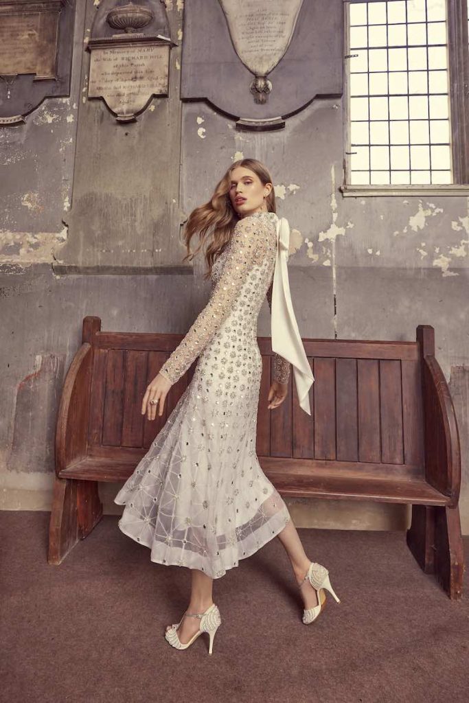 In questa foto una modella cammina indossando un abito da sposa Temperley Bridal 2024 midi al polpaccio con ricami di paillettes e perline all ove, maniche lunghe e fiocco posteriore sulla nuca in raso. La modella indossa anche un paio di sandali open toe abbinati